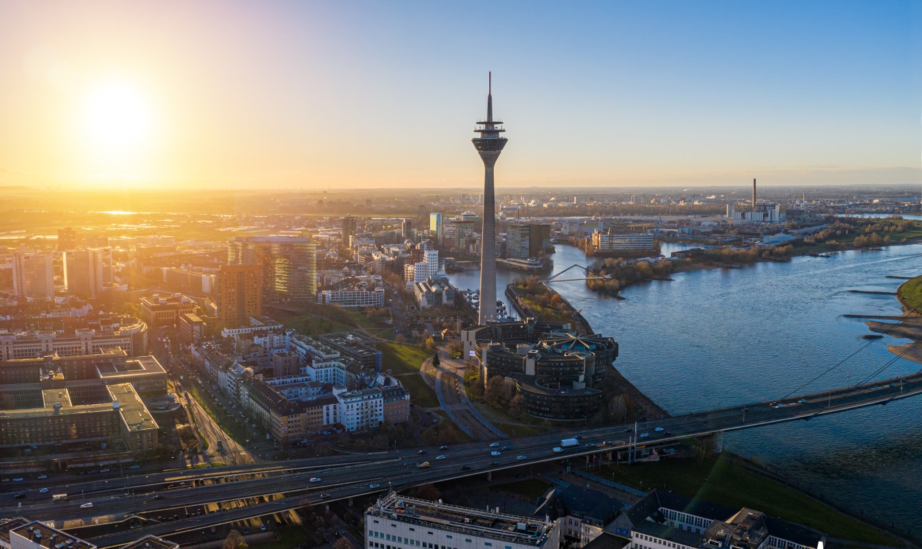 Sonnenaufgang über einer Stadt mit Fluss, einer Hochbrücke und einem markanten Turm.