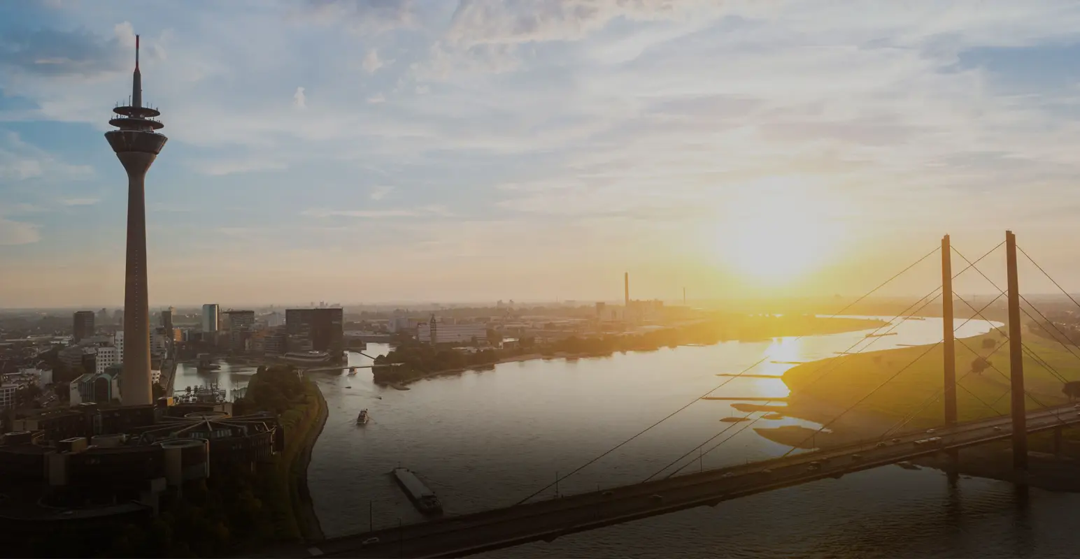Sonnenaufgang über einer Stadt mit Fluss, einer Hochbrücke und einem markanten Turm.