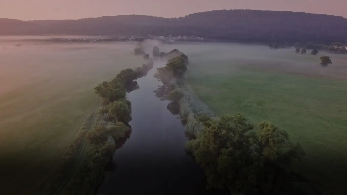 Eine Luftaufnahme zeigt eine Flussbiegung durch eine nebelverhangene Landschaft bei Sonnenaufgang.