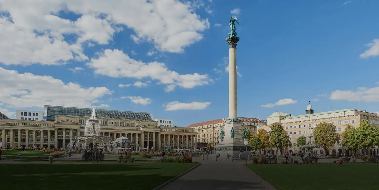 Eine weiträumige Platzansicht mit einem hohen Säulenmonument und einem Springbrunnen, umgeben von Gebäuden und Menschen.