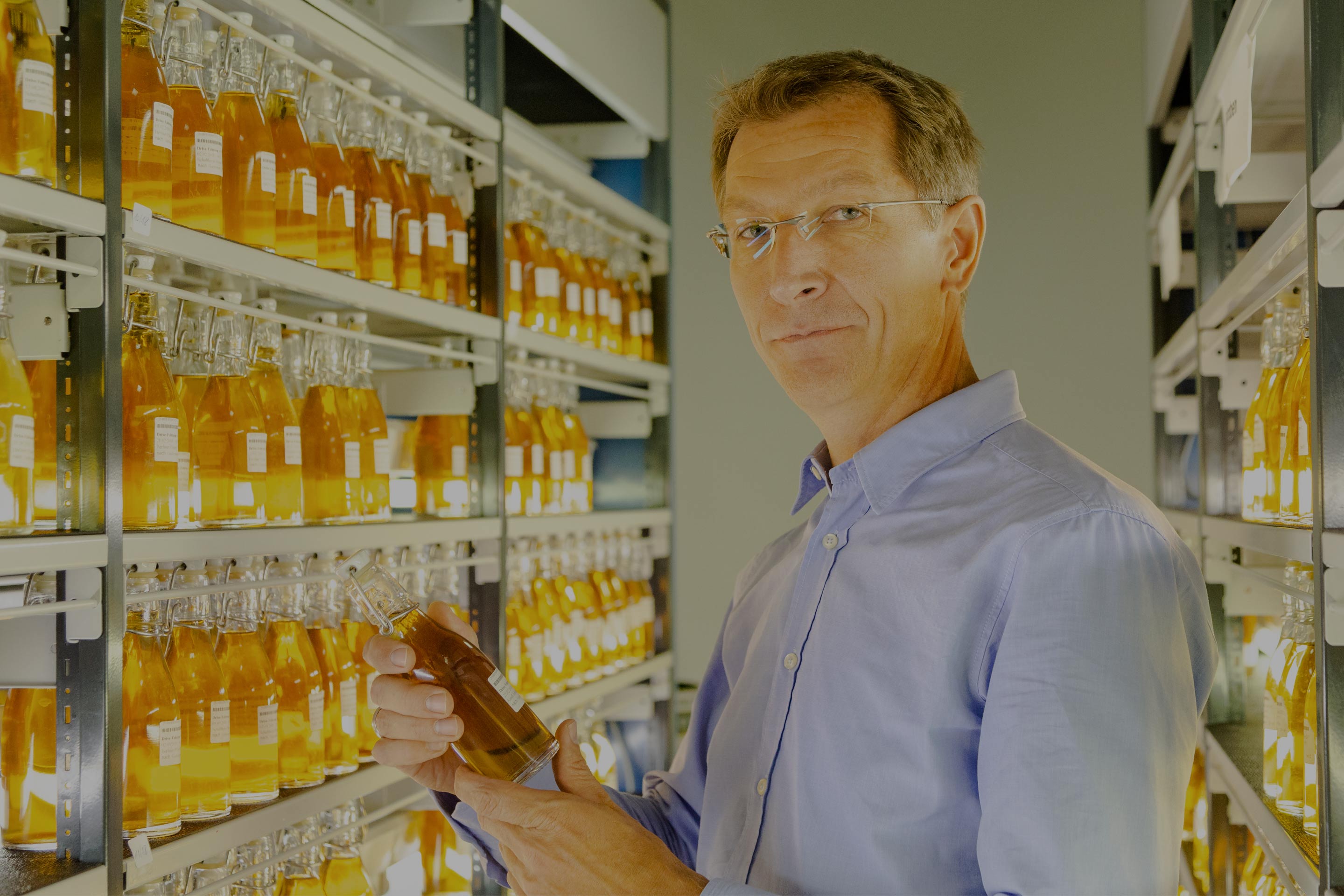 Ein Mann steht vor Regalen mit zahlreichen Flaschen im Labor und hält eine Flasche.