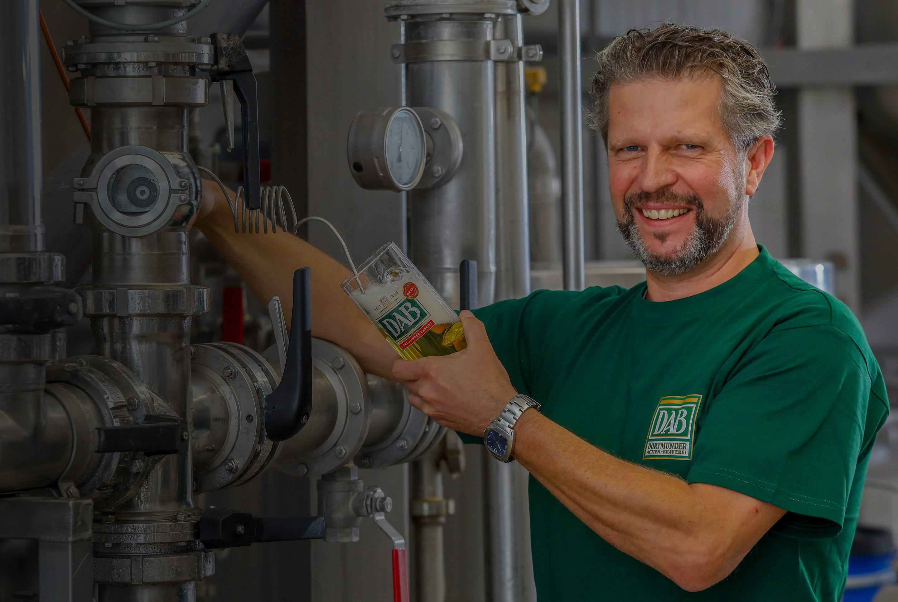 Ein Mann füllt Bier in ein Glas in einer Brauerei. Er lächelt und trägt ein grünes Shirt.