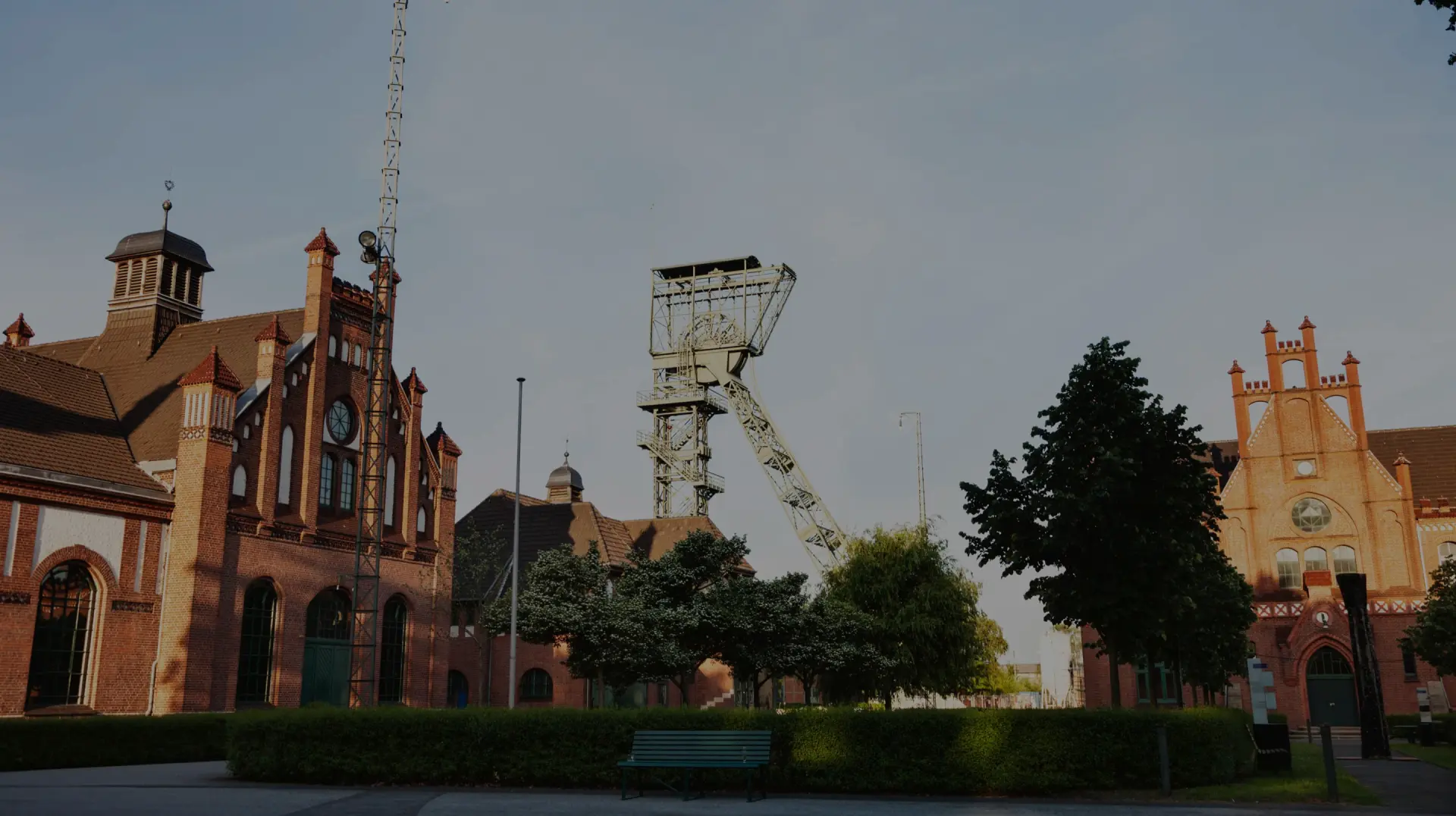 Fördergerüst erhebt sich zwischen historischen Backsteingebäuden. Bäume und Grünflächen umgeben den Platz.