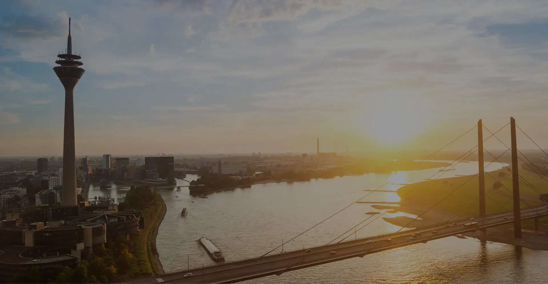 Fernsehturm erhebt sich neben einem Fluss und einer Schrägseilbrücke. Die Sonne steht tief über der Stadtlandschaft.