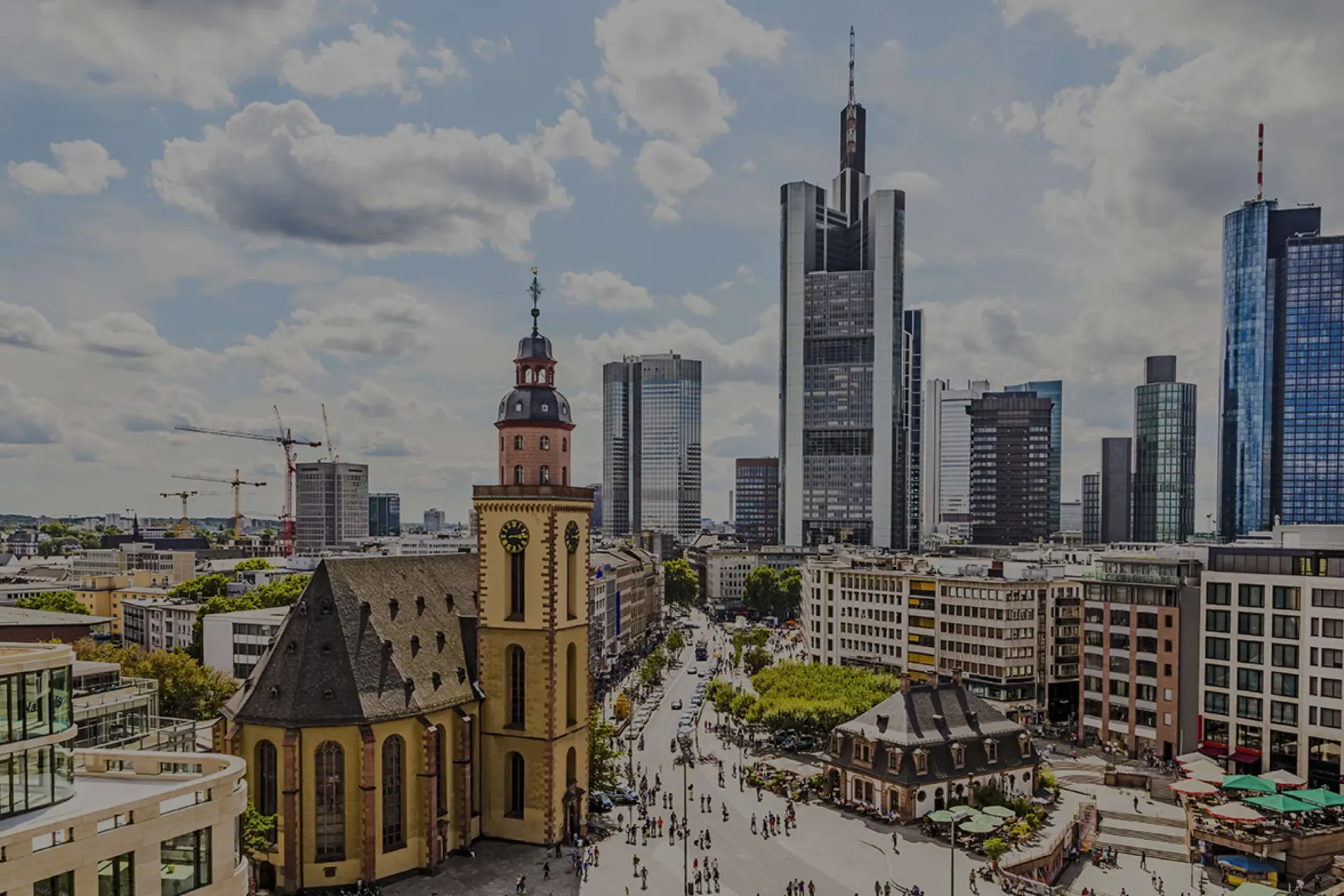 Kirchturm steht vor einer Skyline aus Hochhäusern und Bürogebäuden. Wolken ziehen über die dicht bebaute Stadt.