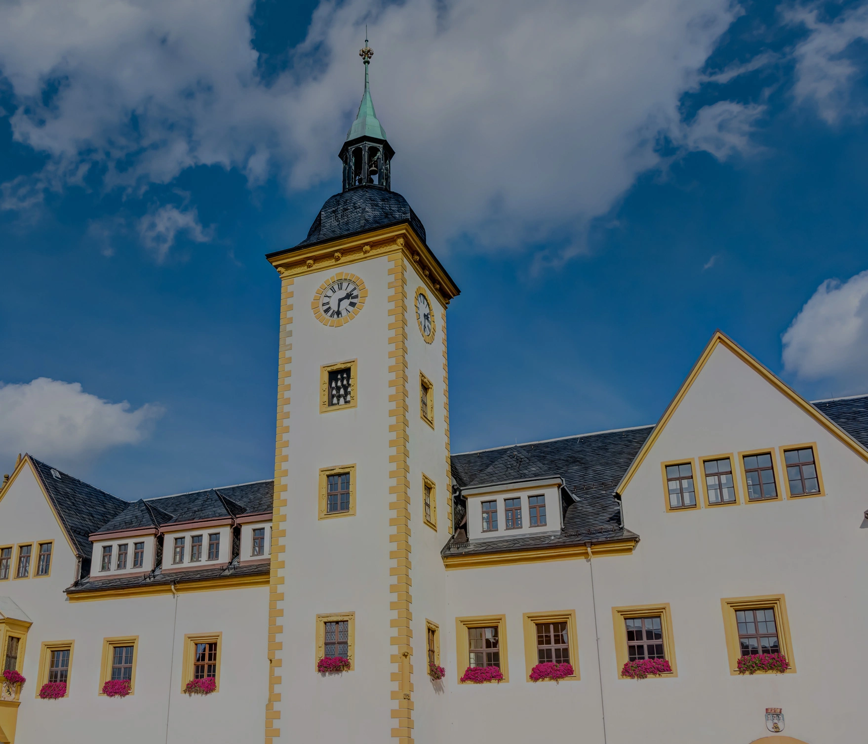 Gebäude mit Uhrturm und Spitzdach prägt einen historischen Gebäudekomplex. Wolken ziehen über den blauen Himmel.