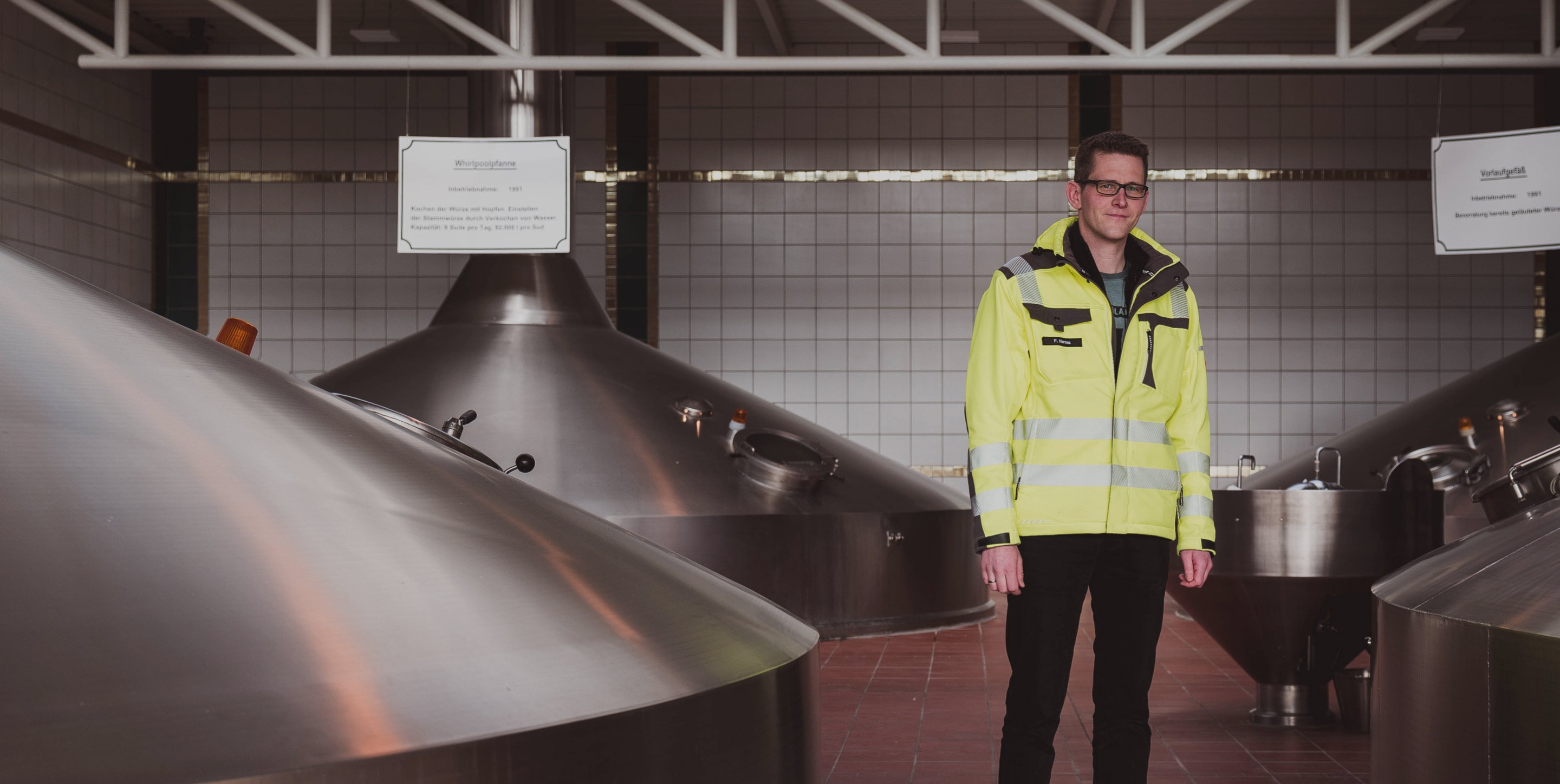 Ein Mann in gelber Sicherheitsjacke steht zwischen großen Braukesseln in einer Brauerei.