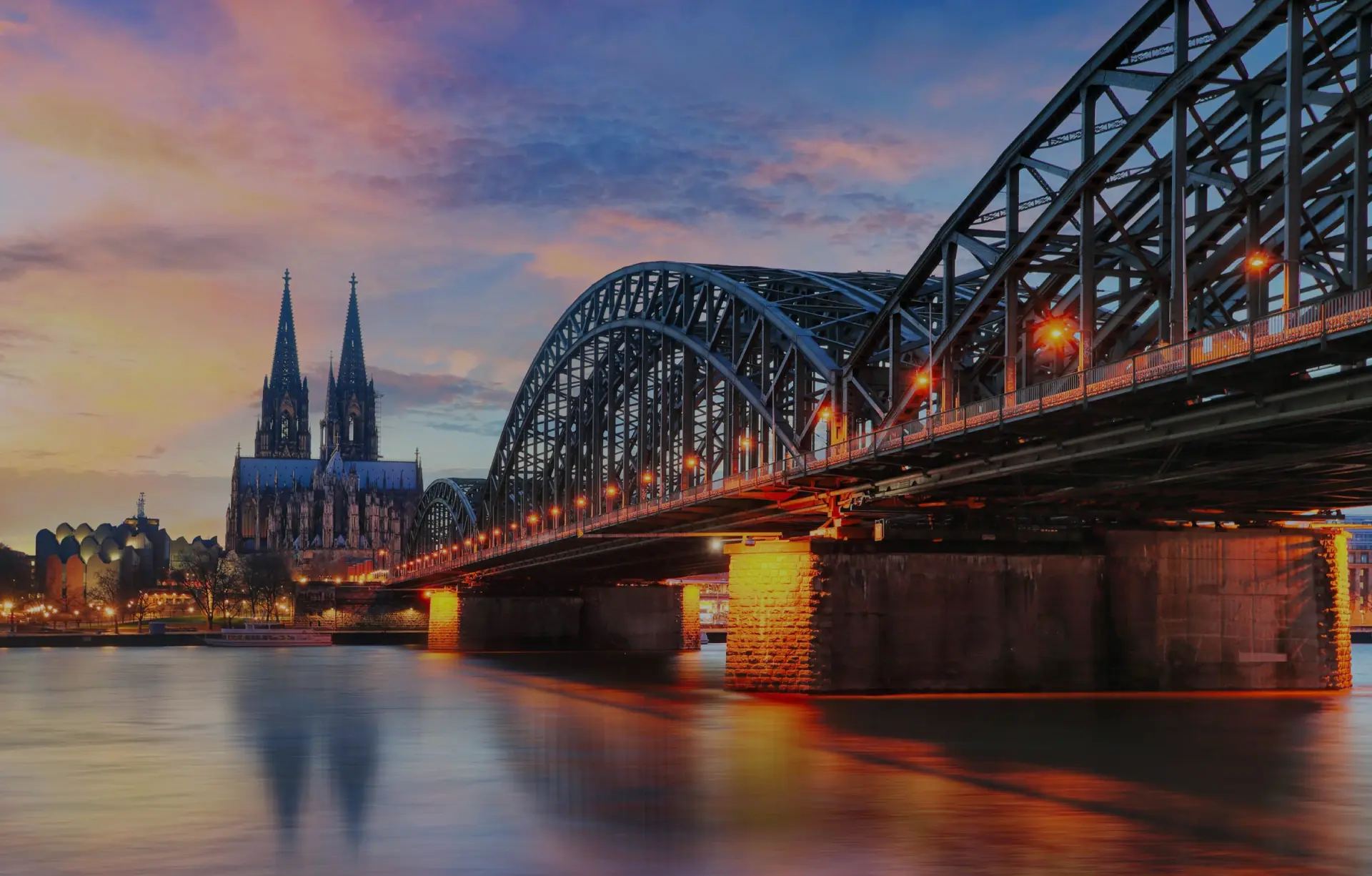 Große Stahlbrücke überspannt einen Fluss vor einer Kathedrale mit Doppeltürmen. Lichter spiegeln sich im Wasser bei Dämmerung.