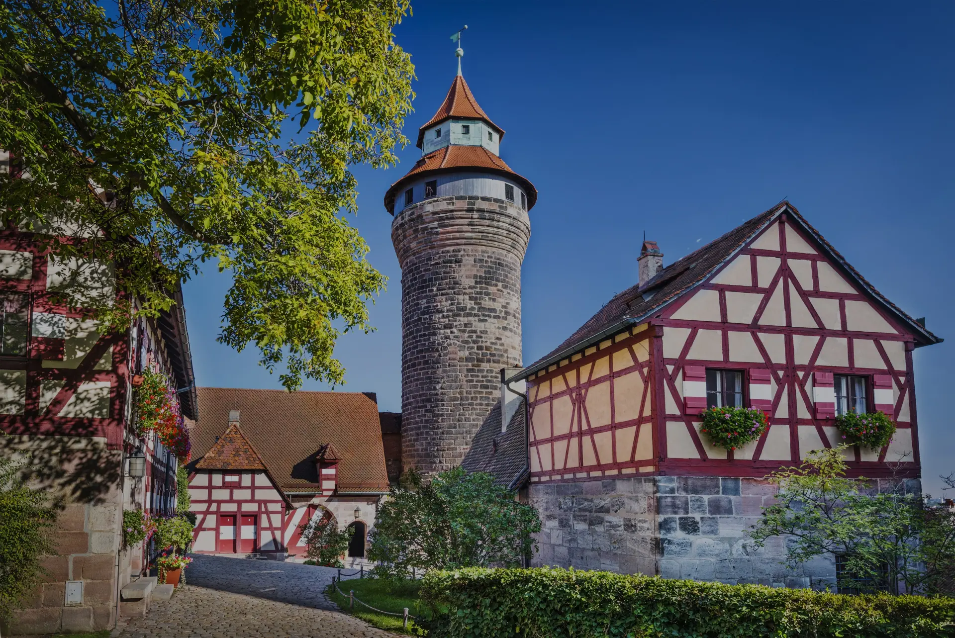 Runder Steinturm erhebt sich zwischen Fachwerkhäusern in einem historischen Ortskern. Bäume und Hecken säumen den Platz.