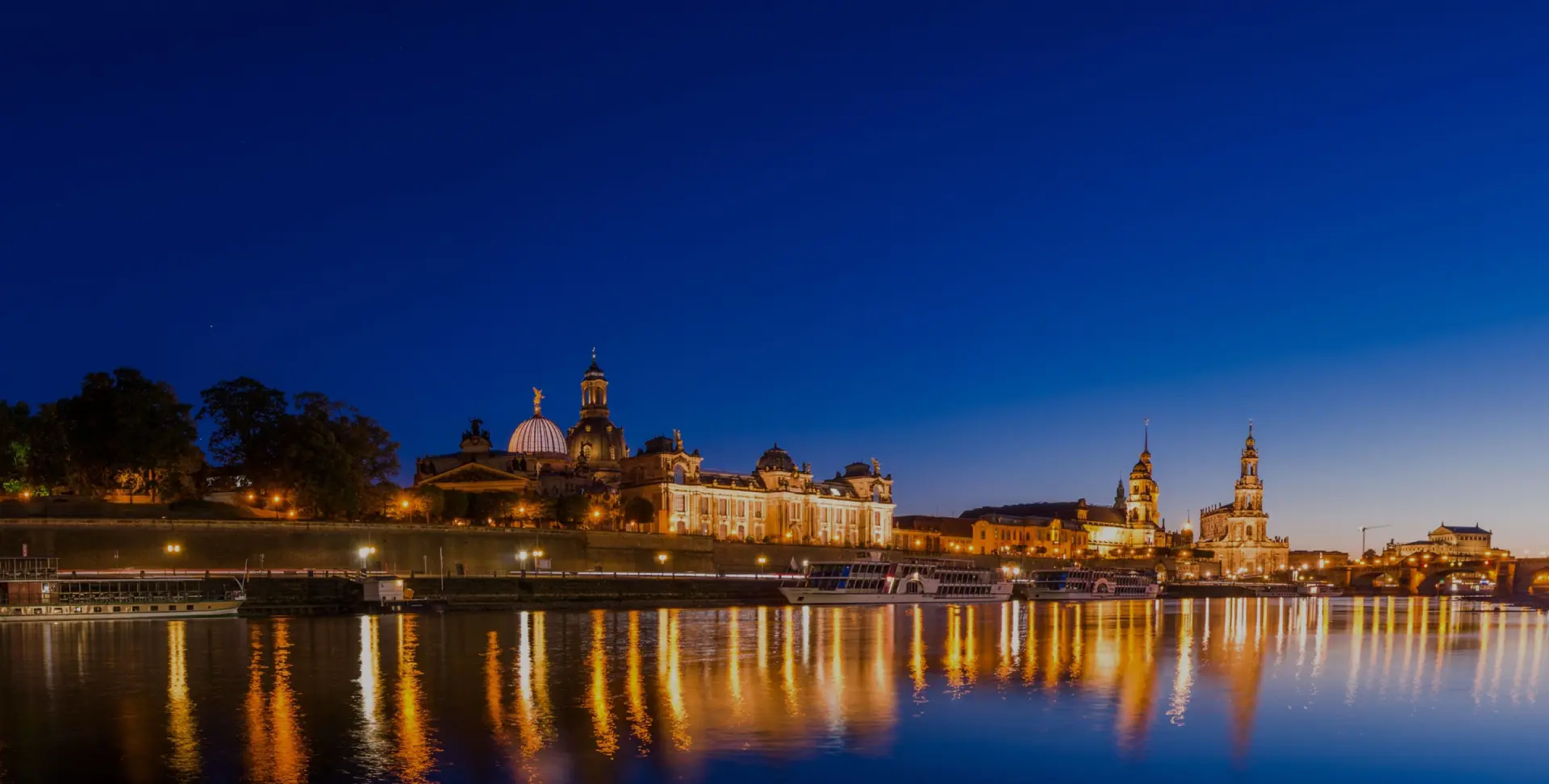 Beleuchtete Gebäude und Kirchtürme stehen am Ufer eines Flusses. Lichter spiegeln sich im Wasser unter dunklem Nachthimmel.