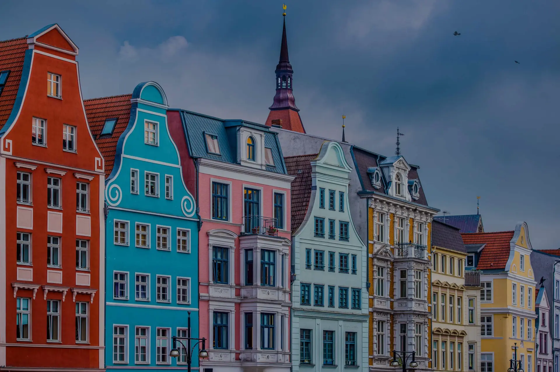 Bunte Giebelhäuser stehen dicht nebeneinander entlang einer Häuserzeile. Kirchturm ragt vor bewölktem Himmel auf.