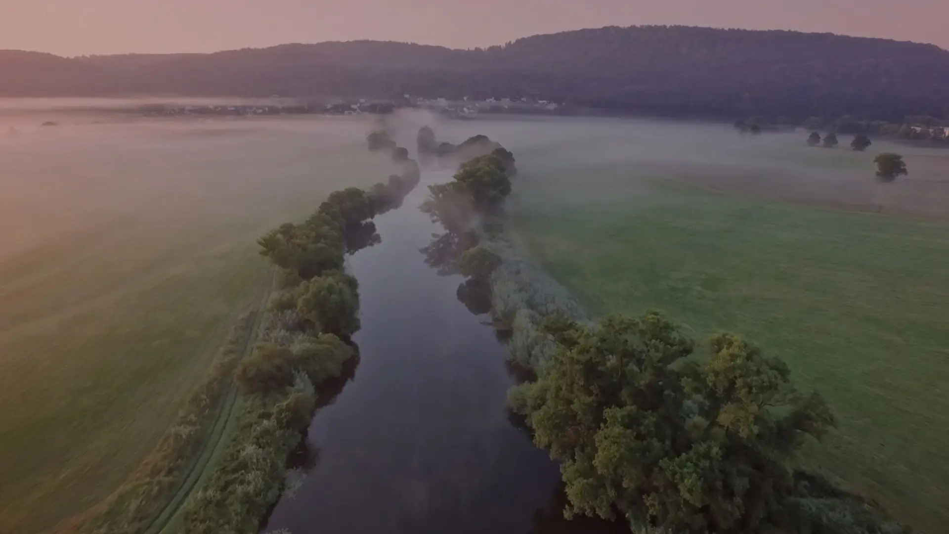 Flusslauf mit Bäumen schlängelt sich durch weite Wiesen. Nebel liegt über der Landschaft vor bewaldeten Hügeln.