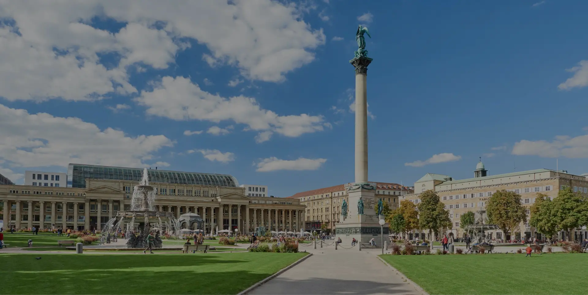 Säule mit Statue auf einem grossen Platz neben einem Springbrunnen. Grünflächen und Gebäude unter blauem Himmel.