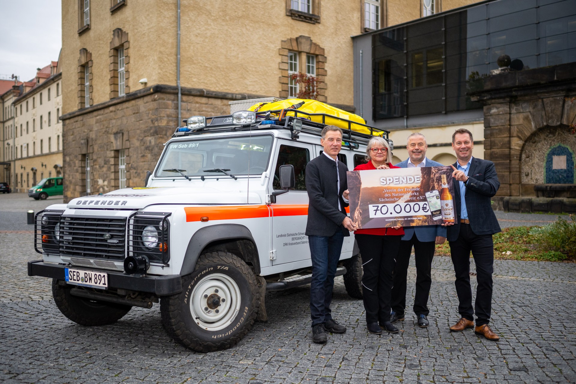Four people are standing in front of an SUV holding a large donation cheque for 70,000 euros.